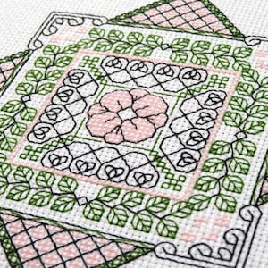 May include: A detailed cross-stitch pattern with a central pink flower, black and white geometric shapes, and green leaf borders. The design is set against a white backdrop, with pink lattice corners.