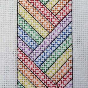 Puede incluir: Un patrón de punto de cruz con un diseño de chevron arcoíris. El patrón está trabajado en hilo negro, blanco, rojo, naranja, amarillo, verde, azul, añil y violeta.