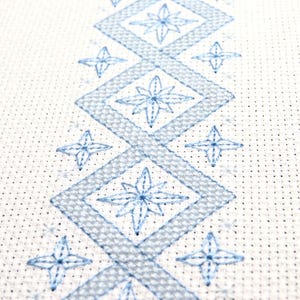 Puede incluir: Primer plano de un patrón de punto de cruz en hilo azul claro sobre tela blanca. El diseño presenta un patrón repetitivo de diamantes y motivos florales en forma de estrella. La tela tiene una textura similar a una cuadrícula, típica de los proyectos de punto de cruz.