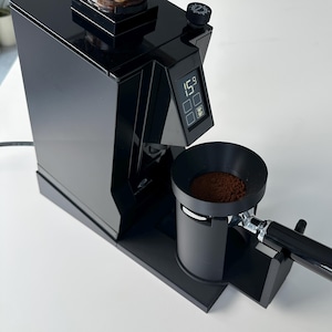 Pode incluir: Um moinho de café preto com um depósito transparente cheio de grãos de café torrados. O dispositivo possui um visor digital que mostra 15.9, um botão de ajuste circular e um suporte de porta-filtro contendo café moído.