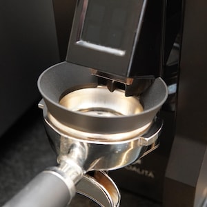 Puede incluir: Primer plano de un molinillo de espresso negro y plateado. El molinillo cuenta con un soporte para portafiltro, una cámara de molienda y una pantalla digital. El portafiltro se inserta en el soporte, listo para recibir café recién molido.