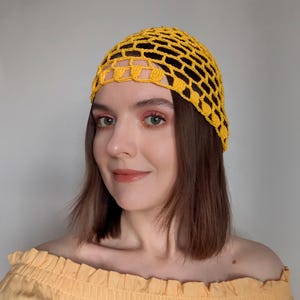 May include: A yellow crocheted head covering with a net-like design. The head covering is worn on the head. The person in the image is wearing a yellow off-the-shoulder top. The background is a solid light grey.
