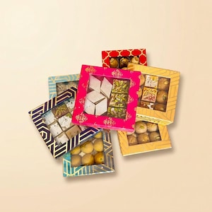 May include: A collection of square gift boxes with clear windows, each containing assorted Indian sweets. The boxes feature vibrant patterns in pink, yellow, blue, and purple, filled with diamond-shaped kaju katli and round laddu confections.