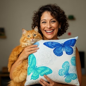 May include: A white square pillow with a blue butterfly design. The pillow features three butterflies in shades of blue and turquoise, with a floral background. The pillow is being held by a person with a ginger cat.