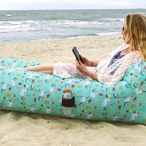 Könnte beinhalten: Ein türkisfarbener aufblasbarer Lounger mit einem Einhorn- und Regenbogenmuster liegt an einem Sandstrand. Eine Wasserflasche befindet sich in einer Seitentasche. Der Lounger ist für Entspannung und den Außenbereich konzipiert.