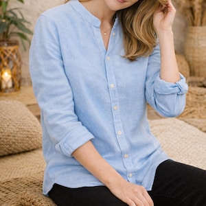 May include: A pale blue linen shirt with a mandarin collar and button-down front. The long sleeves are rolled up. The shirt is paired with black trousers. The model is smiling.