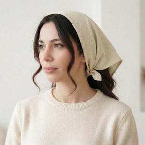 May include: A beige headscarf tied at the nape of the neck. The head covering is made of a soft, lightweight fabric. The person is wearing a cream-colored sweater.
