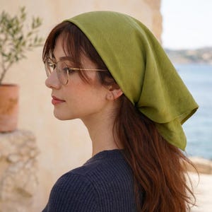 May include: A woman wearing a green headscarf and glasses. The headscarf is a solid, light green color and is tied at the back of the head. The glasses have thin, gold-colored frames. The woman is wearing a dark blue sweater.