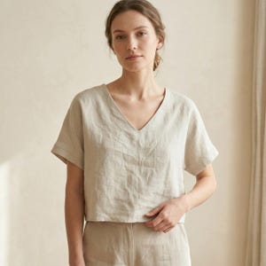 Könnte beinhalten: Ein hellbeige Leinenoberteil mit V-Ausschnitt und kurzen Ärmeln. Das Oberteil wird mit einer passenden beigen Hose kombiniert. Der Stoff wirkt leicht und atmungsaktiv, ideal für warmes Wetter. Das Outfit ist schlicht und elegant.