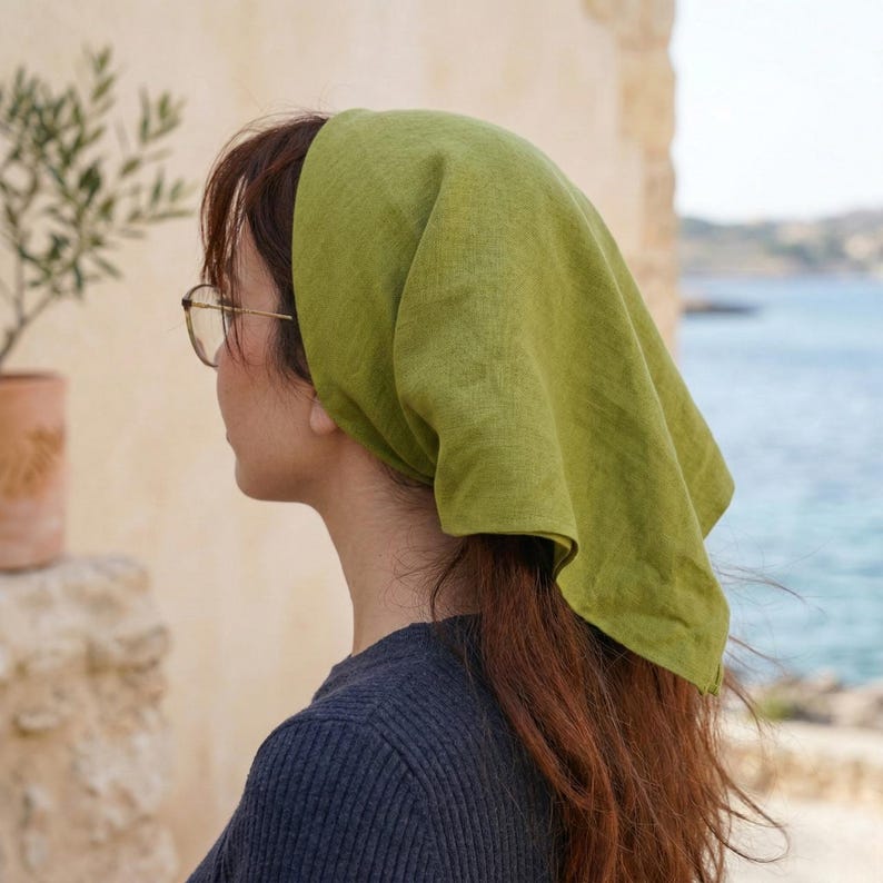 May include: A woman wearing a green headscarf. The headscarf is a solid, light green color and is tied at the back of the head. The woman is wearing glasses and a dark blue sweater. The background includes a stone wall and a view of the ocean.