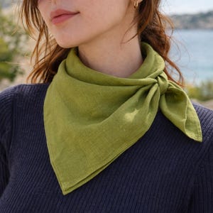 May include: A green linen bandana tied around the neck. The bandana is a vibrant shade of green and is knotted at the front. The person is wearing a dark blue sweater. The background shows a blurred view of the sea and sky.