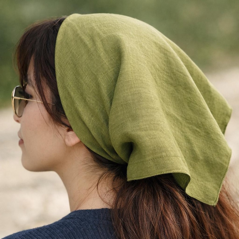 May include: A green linen headscarf is worn on the head. The headscarf is a solid, light green color. The person is wearing sunglasses and a dark blue shirt. The hair is brown and pulled back.