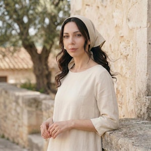May include: A woman wearing a beige headscarf and a long-sleeved, cream-colored dress. The woman is leaning against a stone wall, with a tree and building in the background. The dress has a simple, round neckline.