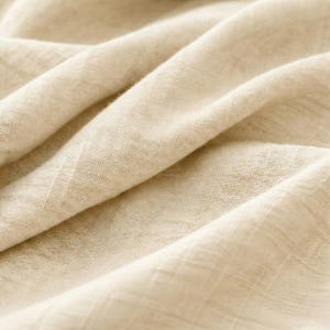 May include: Close-up of a beige fabric with a textured weave. The fabric is folded and draped, creating soft shadows and highlights. The material appears to be lightweight and breathable, suitable for clothing or home decor.