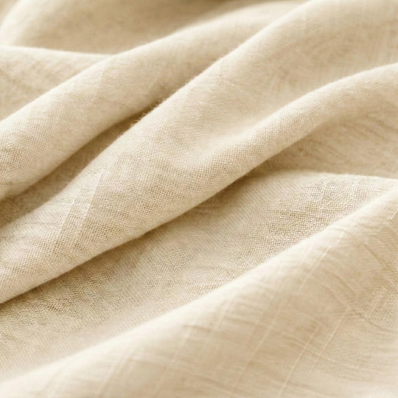 May include: Close-up of a beige fabric with a textured weave. The fabric is folded and draped, creating soft shadows and highlights. The material appears to be lightweight and breathable, suitable for clothing or home decor.
