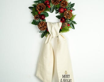 Könnte beinhalten: Ein Weihnachtskranz mit Tannenzapfen, roten Ornamenten und Grünzeug. Ein cremefarbenes Handtuch mit dem Text "MERRY & BRIGHT" hängt darunter. Der Kranz hängt an einer weißen Wand.