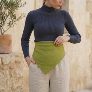 May include: A woman wearing a dark gray ribbed turtleneck sweater, light green square scarf, and beige linen pants. The scarf is wrapped around the waist. The background is a stone wall and a potted plant.