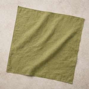 May include: A square, olive green linen napkin. The fabric has a slightly textured appearance with visible weave details. The napkin is folded and lies flat, with soft shadows suggesting its shape. The color is a muted, earthy tone.