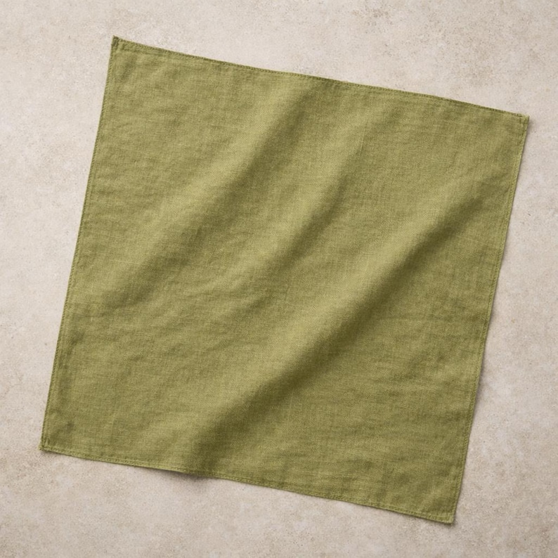 May include: A square, olive green linen napkin. The fabric has a slightly textured appearance with visible weave details. The napkin is folded and lies flat, with soft shadows suggesting its shape. The color is a muted, earthy tone.