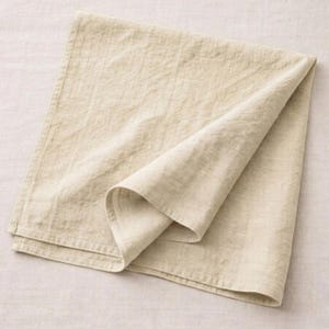 May include: A folded, beige linen napkin. The napkin is rectangular with a folded corner, showcasing the fabric's texture. The neutral color and simple design suggest a focus on natural materials and minimalist aesthetics.
