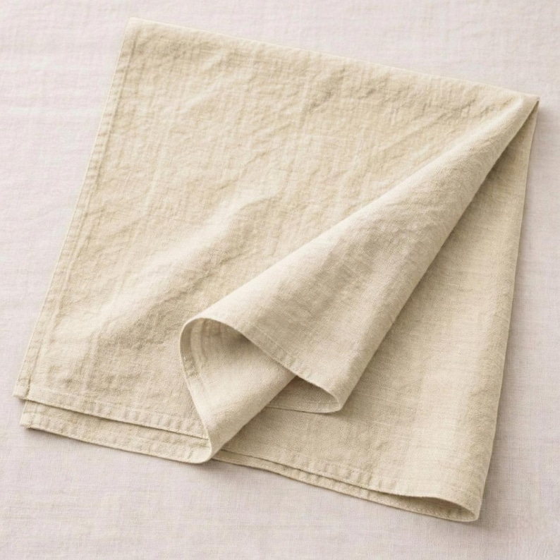May include: A folded, beige linen napkin. The napkin is rectangular with a folded corner, showcasing the fabric's texture. The neutral color and simple design suggest a focus on natural materials and minimalist aesthetics.