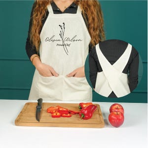 May include: Cream-coloured apron with the text "Olivia Wilson Private Chef" and a botanical illustration. The apron has two front pockets and a cross-back design. A wooden cutting board with a knife, sliced red bell peppers, and whole red apples are in the foreground.