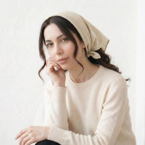 May include: A woman wearing a beige headscarf and a cream-colored sweater. The headscarf is tied at the back of the head. The sweater has a ribbed crew neck and long sleeves. The woman is looking at the camera.
