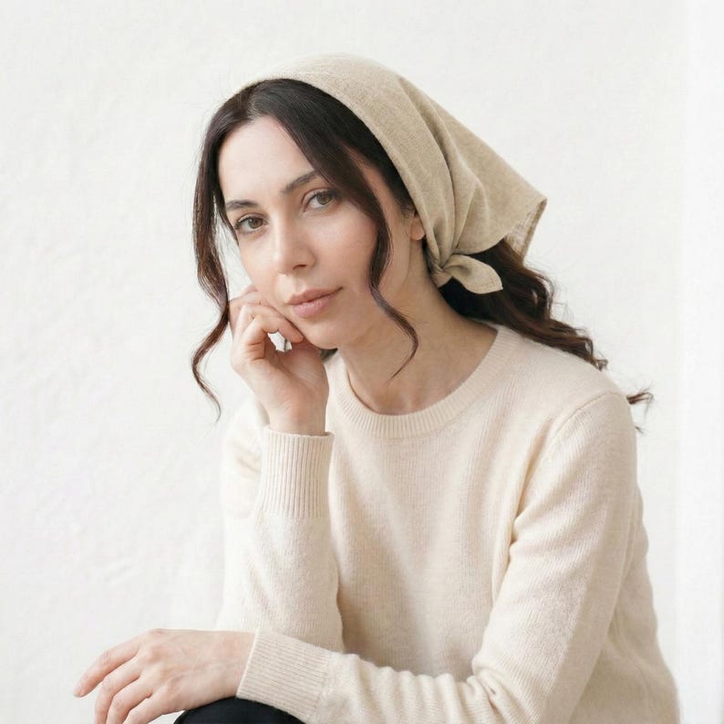 May include: A woman wearing a beige headscarf and a cream-colored sweater. The headscarf is tied at the back of the head. The sweater has a ribbed crew neck and long sleeves. The woman is looking at the camera.