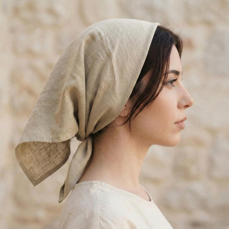 May include: A beige linen headscarf tied at the nape of the neck. The head covering is draped over the head and tied in a knot at the back. The fabric has a slightly textured appearance. The person is wearing a light-colored top.