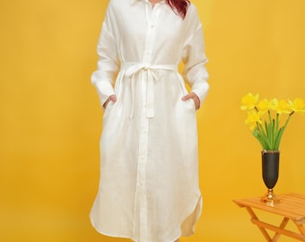 May include: A long, off-white linen shirt dress with long sleeves, a button-down front, and a matching tie belt. The dress has pockets and a midi length, and is shown against a yellow background.