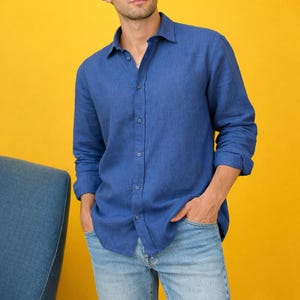 May include: A blue button-down shirt with long sleeves. The shirt is made of a textured fabric and is paired with light blue jeans. The background is a solid yellow color. The shirt has a classic collar and buttoned cuffs.