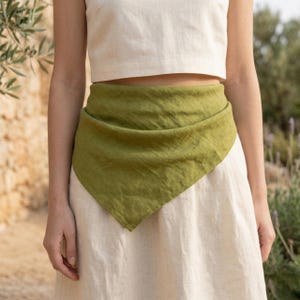 May include: A woman wearing a white crop top and skirt with a green scarf wrapped around the waist. The scarf is a solid olive green color and is made of a soft, flowing fabric. The outfit is simple and elegant.