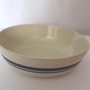 May include: Cream-colored ceramic bowl with two navy blue stripes around the exterior. The bowl has a rounded shape and a glossy finish. This kitchenware item is suitable for serving food.