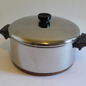 May include: A stainless steel cooking pot with a matching lid, featuring two black side handles and a black knob on the lid. The pot has a polished metallic finish and a copper-colored base, presenting a cylindrical shape for kitchen use.