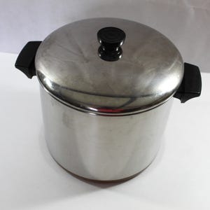 May include: Stainless steel cooking pot with a lid and black handles. The pot has a shiny, metallic finish and a black knob on the lid. The pot is designed for cooking and food preparation.