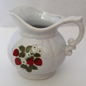 May include: A pearlescent white ceramic pitcher with a rounded body and a curved handle. The pitcher features a strawberry and flower design in red, green, and white. The pitcher has a spout for easy pouring.