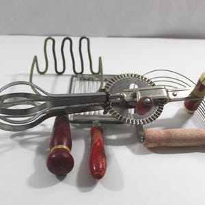 May include: Vintage kitchen tools including a hand-crank egg beater, a pie cutter, and a wire rack. The tools have wooden handles with red and gold accents. The metal components are silver.