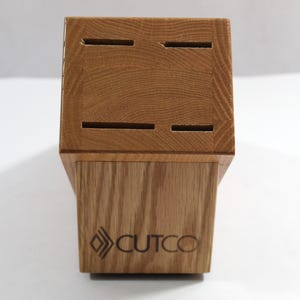 May include: A wooden knife block with slots for knife storage. The block is made of light brown wood and features the "CUTCO" logo on the front. The block has a rectangular shape with a slightly angled base.