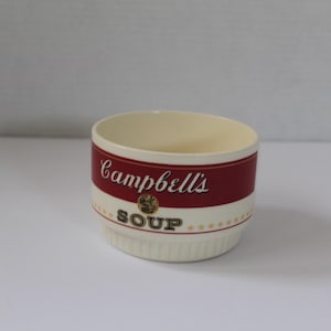 May include: Cream-colored ceramic soup bowl with a red band featuring the "Campbell's Soup" logo. The bowl has a classic design with a cream-colored interior and a white base with a series of small ridges.