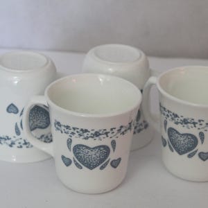 May include: Four white ceramic mugs with a blue heart design. The mugs have a handle and a decorative border of blue leaves. One mug is upside down, revealing the bottom.