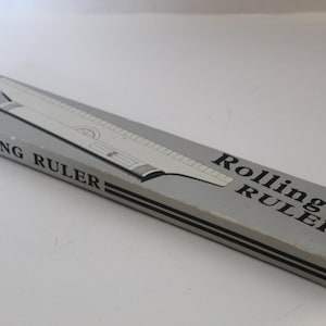 May include: A gray rectangular box with the words "Rolling Ruler" printed on it. The box contains a rolling ruler, a drafting tool. The ruler is silver and white, with a black stripe on the box.