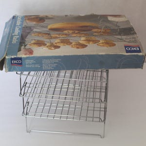 May include: Stackable cooling racks with a box on top. The box is blue and white with images of baked goods. The racks are made of shiny, silver-colored metal. The box reads "EKCO" and "Stackable Cooling Racks."