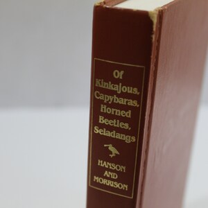 May include: A hardcover book with a reddish-brown cover. The spine features gold lettering that reads "Of Kinkajous, Capybaras, Horned Beetles, Seladangs." The author's name, Hanson and Morrison, is also in gold. A small bird illustration is present.
