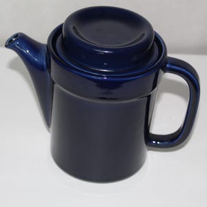 May include: A navy blue ceramic teapot with a matching lid and handle. The teapot has a spout and a rounded body. The lid sits on top of the teapot. The teapot is a solid color and has a glossy finish.