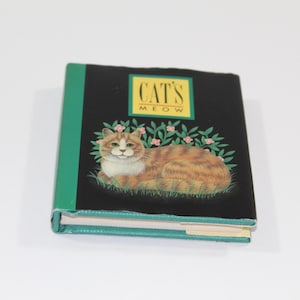 May include: A small book with a black cover featuring a cat illustration. The book's title, "CAT'S MEOW," is in a yellow box. The cat is orange and white, lying on green grass with pink flowers. The spine is green.