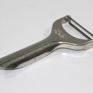 May include: A silver-colored vegetable peeler with a curved handle and a blade. The peeler has a rounded tip and a curved blade guard. The handle appears to be made of metal. The peeler is designed for kitchen use.