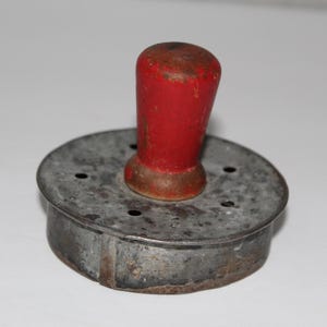 May include: A vintage, round cookie cutter with a red wooden handle. The metal cutter has a circular shape with several small holes and a slightly rusted appearance, suggesting age and use. The handle is cylindrical.
