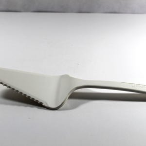 May include: A white plastic cake server with a serrated edge and a curved handle. The handle has a small hole at the end for hanging. This kitchen utensil is designed for serving cakes and pies.