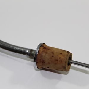 May include: A wine bottle stopper with a natural cork and a silver-colored metal handle. The stopper has a curved design with a small, flat disc at the end. The cork is cylindrical with a metal ring around it.