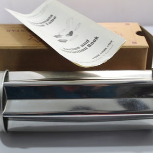 May include: A stainless steel pasta maker with a recipe and instruction book. The pasta maker is cylindrical with a ridged design. The box is brown with a star logo.